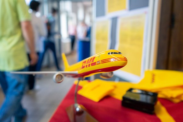 Im Vordergrund ist ein gelbes Flugzeugmodell des Logistik-Unternehmens DHL zu sehen. Im Hintergrund lässt sich ein Messestand des Unternehmens erahnen.