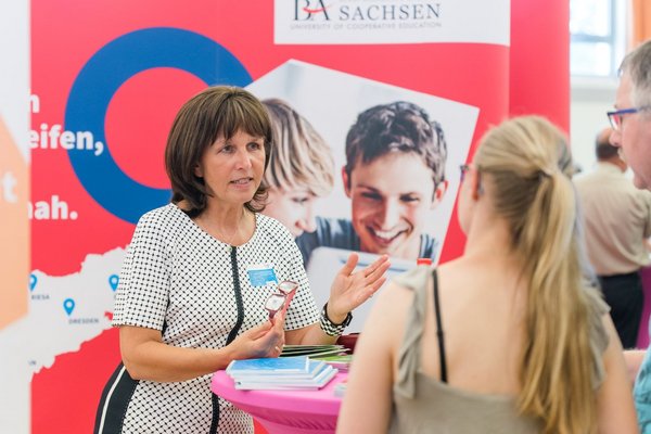 Eine Beraterin der Dualen Hochschule Sachsen spricht mit zwei jungen Messebesuchern an einem Infostand. Der Stand ist in Rot und Blau gestaltet, mit einem großen Werbebanner im Hintergrund, das die Duale Hochschule Sachsen bewirbt. Auf dem Tisch liegen Info-Broschüren und Materialien zur Studienberatung.