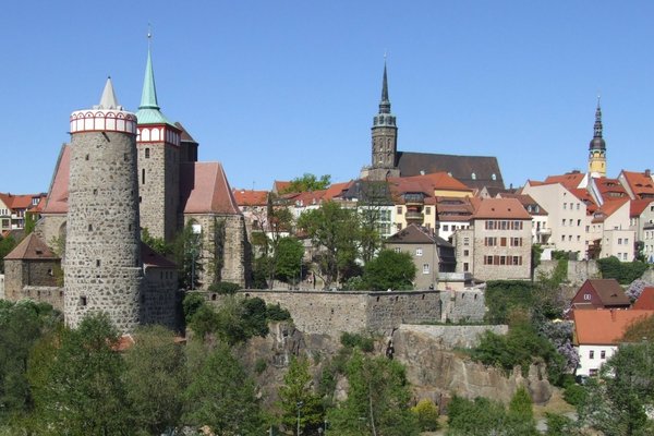 Panoramablick auf die Altstadt von Bautzen mit historischen Gebäuden, Türmen und einer Stadtmauer, umgeben von grüner Natur und klarem blauen Himmel.