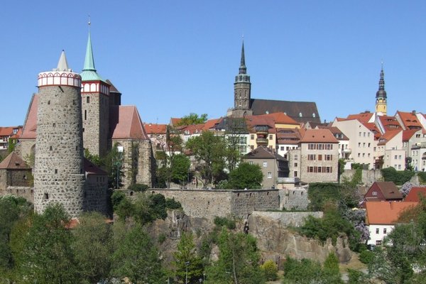 Panoramablick auf die Altstadt von Bautzen mit historischen Gebäuden, Türmen und einer Stadtmauer, umgeben von grüner Natur und klarem blauen Himmel.