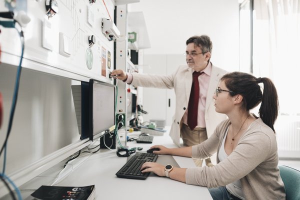 Ein Laborleiter mit weißem Kittel kontrolliert einen Versuchsaufbau. Eine Frau sitzt im Vordergrund und arbeitet an einem Computer in einer Laborumgebung.