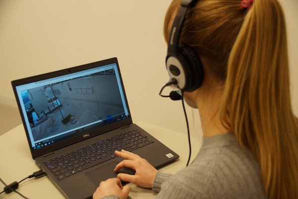 Eine Frau sitzt mit einem Headset auf dem Kopf vor einem Laptop und schaut sich ein Video zu einem Laborversuch an. In dem Bild vom Video ist ein Labor zu sehen. Auf dem Tisch stehen Glasgefäße und ein Arm und eine Hand mit weißem Handschuh einer Person in weißem Kittel sind zu sehen.