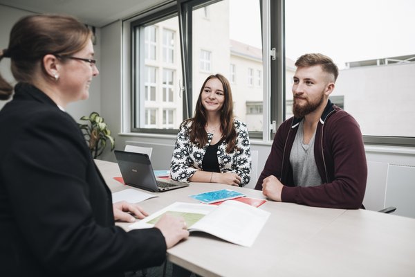 Zwei Studierende sitzen an einem Tisch gegenüber einer Studienberaterin in einem modernen, hellen Büro. Auf dem Tisch liegen Unterlagen und ein Laptop. Durch die Fenster ist der Blick auf Gebäude im Hintergrund sichtbar.