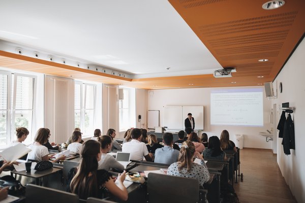 Eine Gruppe von Studierenden sitzt in einem modernen Hörsaal mit Holz- und Weißakzenten. Sie hören einem Vortragenden zu, der vorne vor einer Projektionsleinwand steht und eine Präsentation hält. Tageslicht strömt durch die großen Fenster auf der linken Seite.