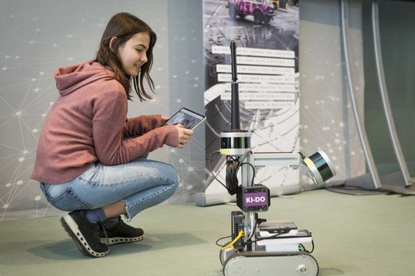 Auf dem Bild ist ein hockendes Mädchen zu sehen, welches ein Tablet hält. Neben dem Mädchen steht ein Roboter.